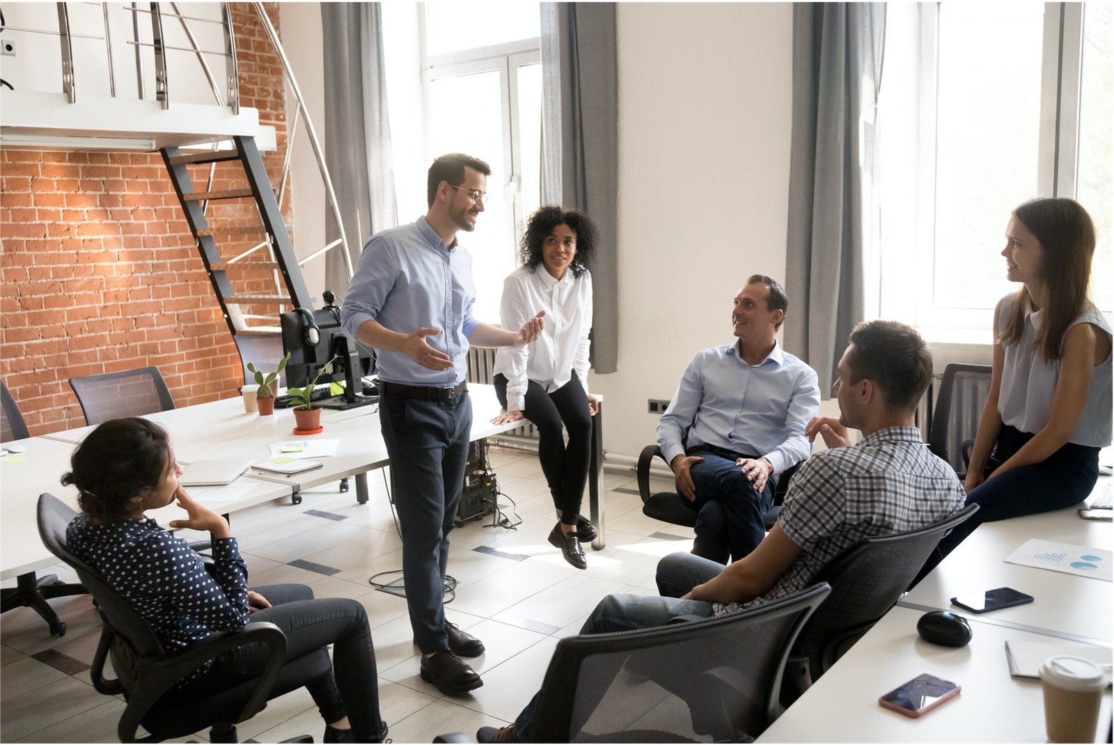 A group of men and women having an informal meeting in an office. A man stands in the centre of the group while speaking, the others are seated as they listen and smile.